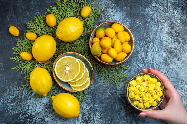 Free photo overhead view of collection of cut and whole natural organic fresh citrus fruits on fir branches and hand holding candies in a small pot on dark background
