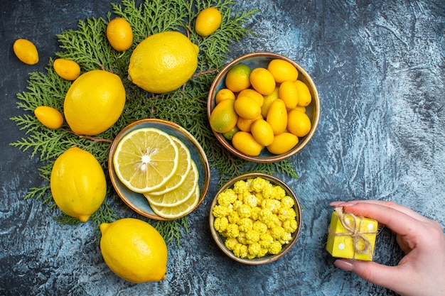 Overhead view of collection of cut and whole natural organic fresh citrus fruits on fir branches and a pot with candies hand holding a gift box on dark background