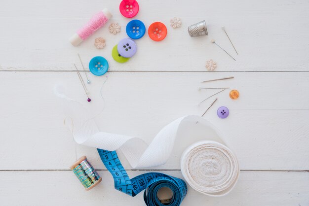 An overhead view of colorful buttons; thimble; needles; ribbon and measuring tape on white wooden desk