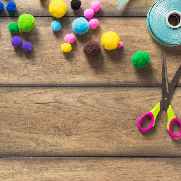 An overhead view of colorful cotton ball; ribbon and scissor on wooden plank