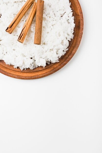 An overhead view of cooked rice with cinnamon sticks on wooden plate against white background