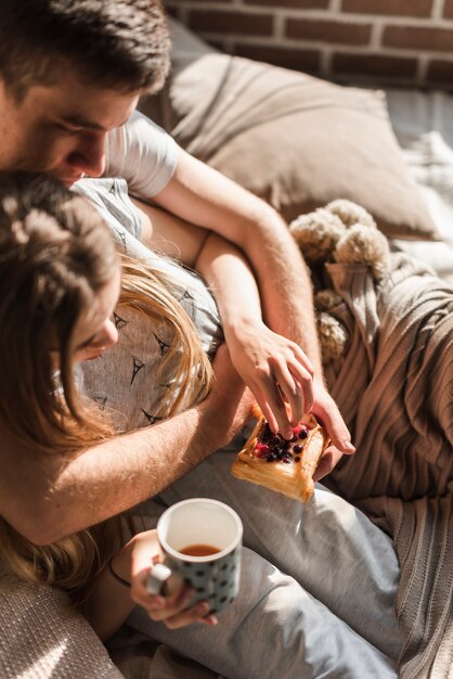 An overhead view of couple lying on bed holding pastry with berries and coffee cup