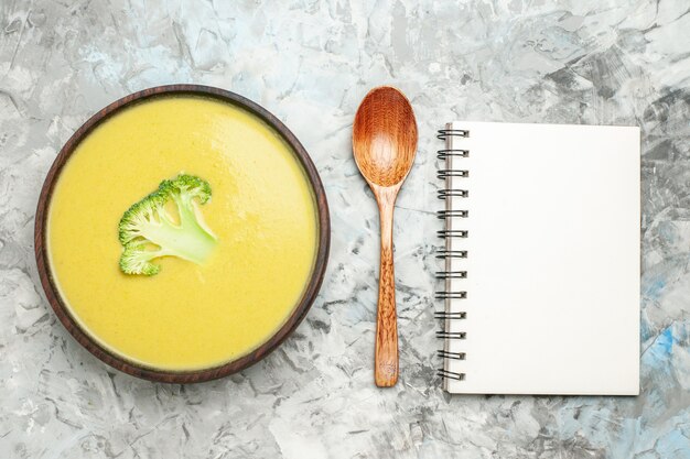 Overhead view of creamy broccoli soup in a brown bowl and spoon next to notebook on gray table