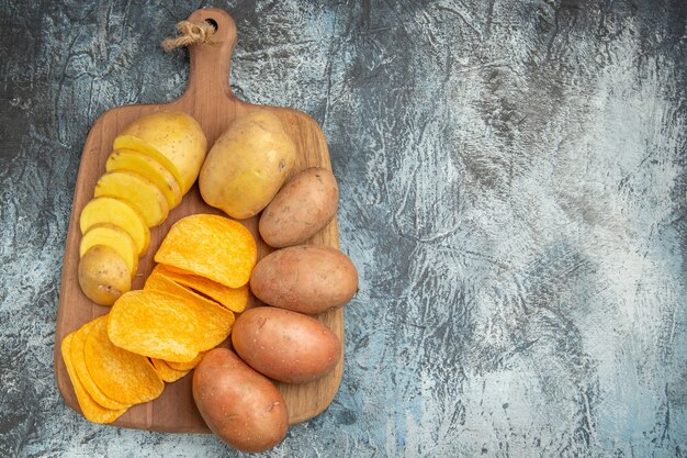 Overhead view of crispy chips and uncooked potatoes on wooden cutting board on gray table