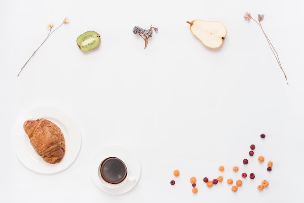 An overhead view of croissant coffee halved kiwi; pears; dried flower and raspberries on white background