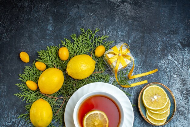 Overhead view of a cup of black tea with lemon and collection of half whole citrus fruits on fir branches and candies in a pot on dark background