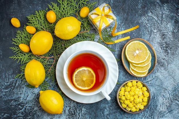 Overhead view of a cup of black tea with lemon and collection of natural organic citrus fruits on fir branches and candies in a pot on dark background