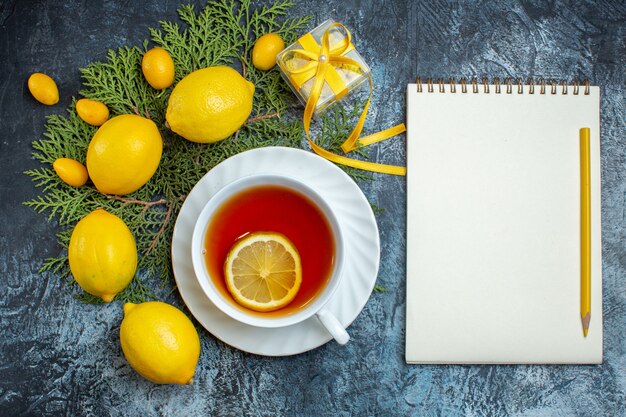 Vista dall'alto di una tazza di tè nero al limone e raccolta di agrumi freschi biologici naturali su rami di abete accanto al taccuino con penna su sfondo scuro Foto Gratuite