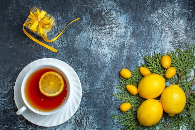 Overhead view of a cup of black tea with lemon and yellow gift box next to citrus fruits collection on fir branches on dark background