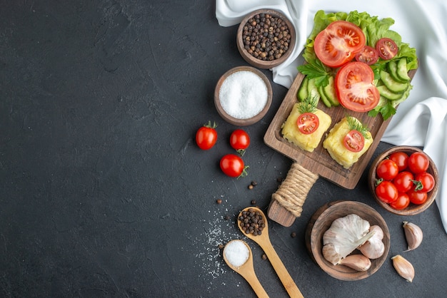 Overhead view of cut whole fresh tomatoes and cucumbers cheese on wooden board cutlery set spices in spoons on black surface