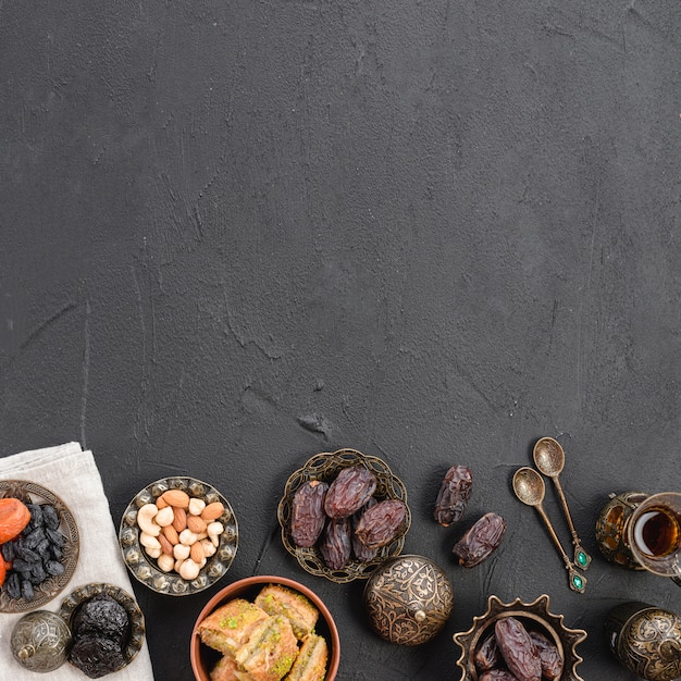 An overhead view of dates; nuts and baklava metallic plates on black concrete textured backdrop with copy space