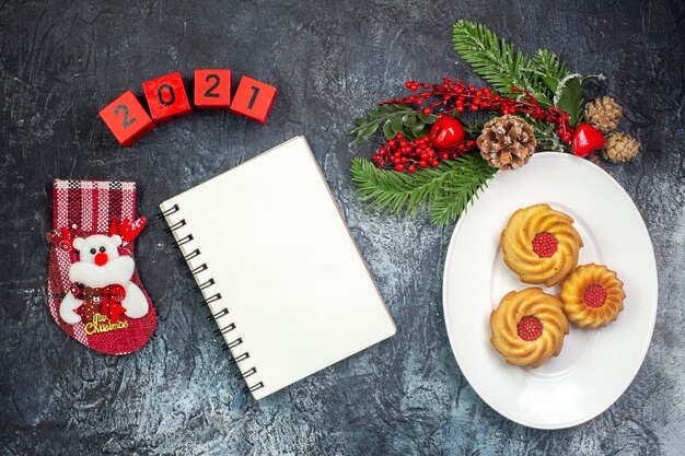 Overhead view of delicious biscuits on a white plate and decorations santa claus hat numbers new year sock next to notebook on dark surface