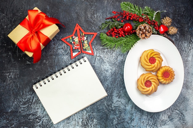 Overhead view of delicious biscuits on a white plate and new year decorations gift with red ribbon next to notebook on dark surface