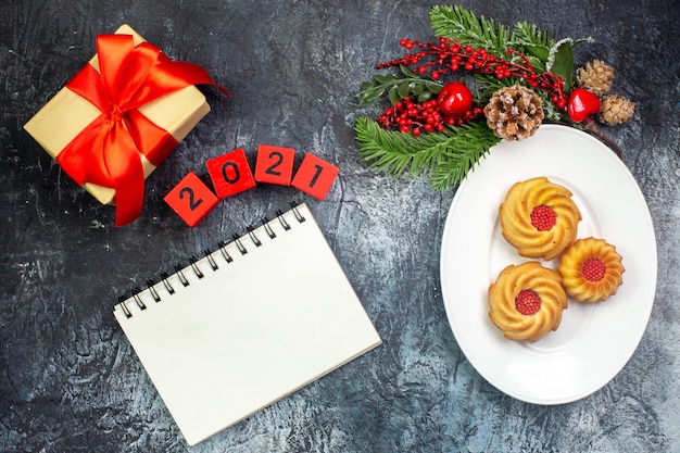 Overhead view of delicious biscuits on a white plate and new year decorations gift with red ribbon next to notebook numbers on dark surface