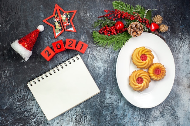 Vista dall'alto di deliziosi biscotti su un piatto bianco e decorazioni di capodanno cappello di babbo natale accanto ai numeri del taccuino su superficie scura