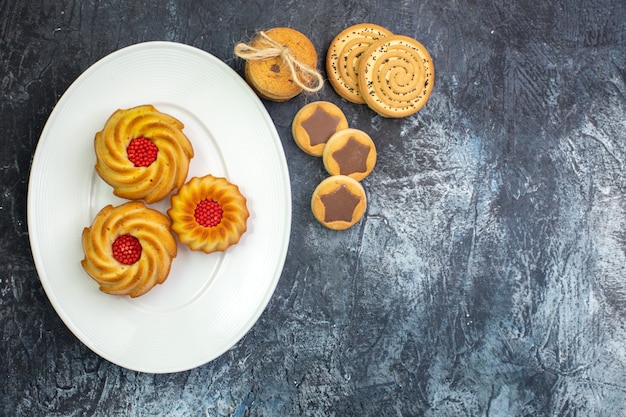 Overhead view of delicious biscuits on a white plate and various cookies and fork on dark surface