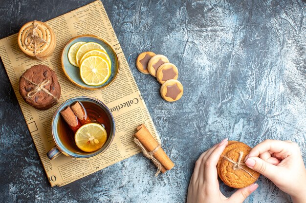 暗い背景に古い新聞にシナモン入りの紅茶のカップを持つおいしいクッキーと手の俯瞰