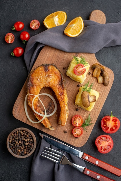 Overhead view of delicious fried fish meal with mushrooms tomatoes cheese on wooden board lemon slices pepper on dark color towel cutlery set on black surface