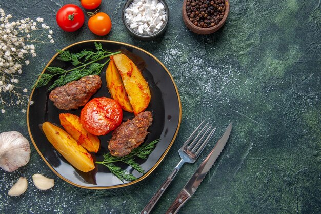Overhead view of delicious meat cutlets baked with potatoes and tomatoes on a black plate cutlery set white flowers spices garlics on green black mixed colors background