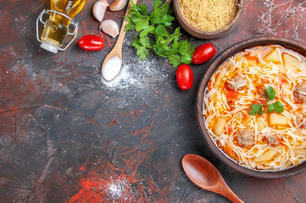 Overhead view of delicious noodle soup with chicken and uncooked pasta in a small brown bowl and spoon garlic tomatoes and greens on the dark background