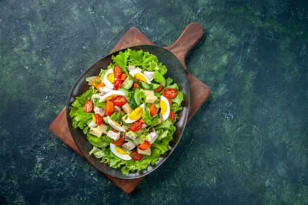 Overhead view of delicious salad with many fresh ingredients on wooden cutting board on black green mix colors background