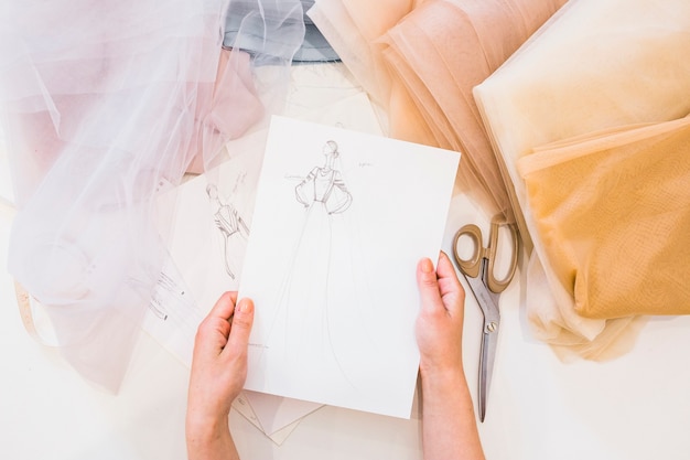 Overhead view of designer's hand holding fashion sketch over workdesk