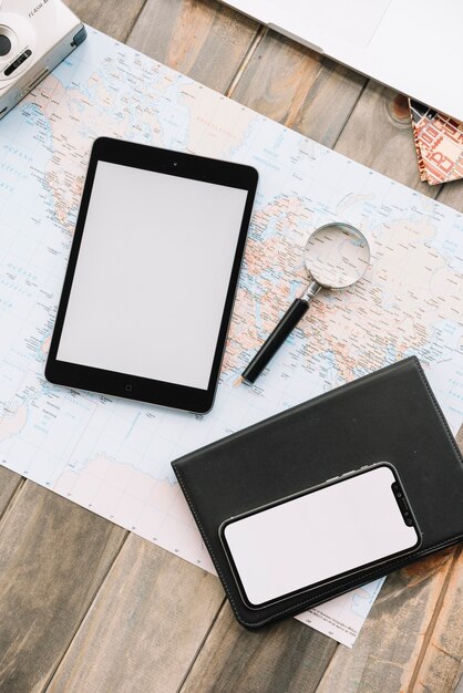 An overhead view of digital tablet; cell phone; magnifying glass and diary on map against wooden backdrop