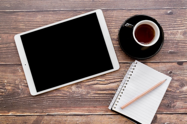 An overhead view of digital tablet; coffee cup and spiral notebook with pencil on wooden textured table