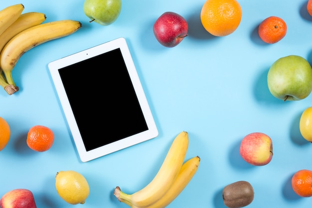 An overhead view of digital tablet with black screen surrounded with colorful fruits on blue background