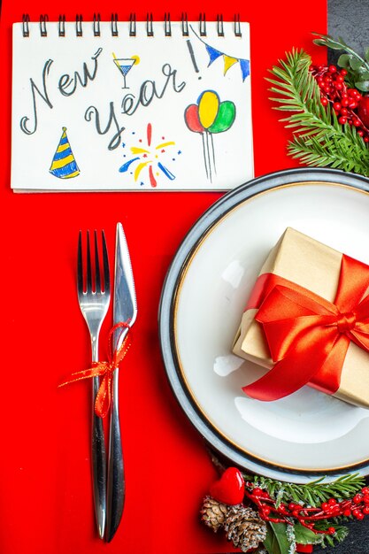 Вид сверху на обеденные тарелки с подарком и еловые ветки, набор столовых приборов, украшение, аксессуар, хвойная шишка, следующая записная книжка с новогодним письмом и рисунками на красной салфетке