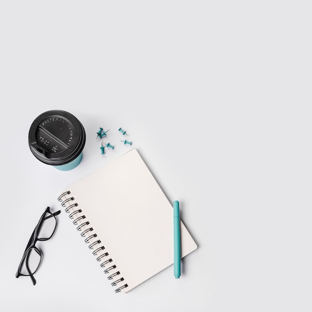 An overhead view of disposable coffee cup; push pins; pen; eyeglasses and spiral notepad on white background