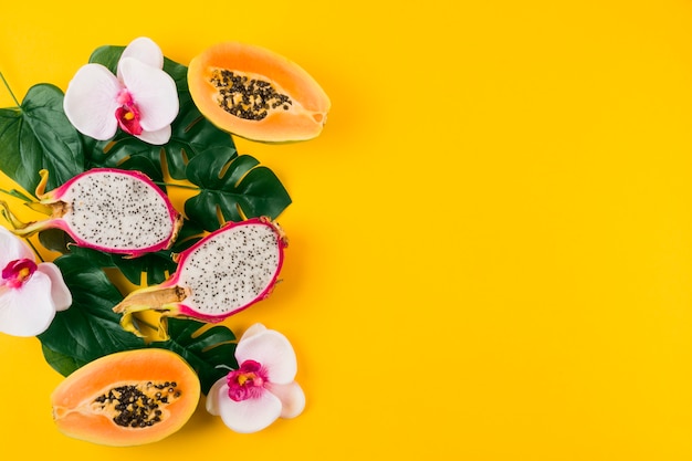 An overhead view of dragon fruits; halved papaya with leaves and orchid flower on yellow backdrop