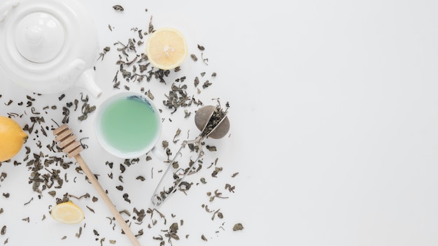 An overhead view of dry tea leaves; tea strainer; lemon; green tea; honey dipper and teapot on white backdrop