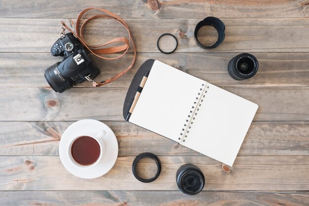 Overhead view of dslr camera; cup of tea; spiral notepad; pen; camera lens and extension rings on wooden background