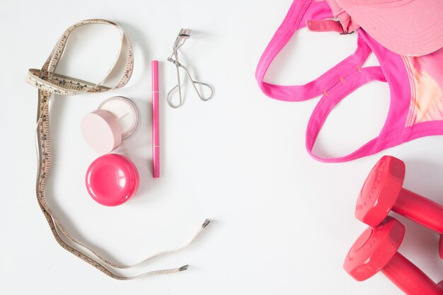 Overhead view of essential beauty items, red dumbbells, sport bra and cosmetic, top view isolated on white background