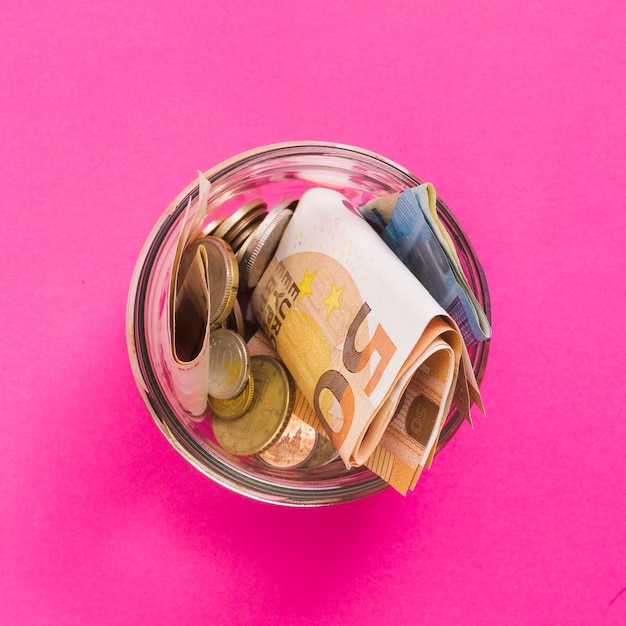An overhead view of euro banknotes and coins in open glass jar against pink background