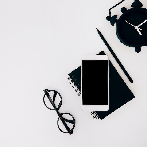 An overhead view of eyeglasses; cellphone; diary; pencil and alarm clock on white background