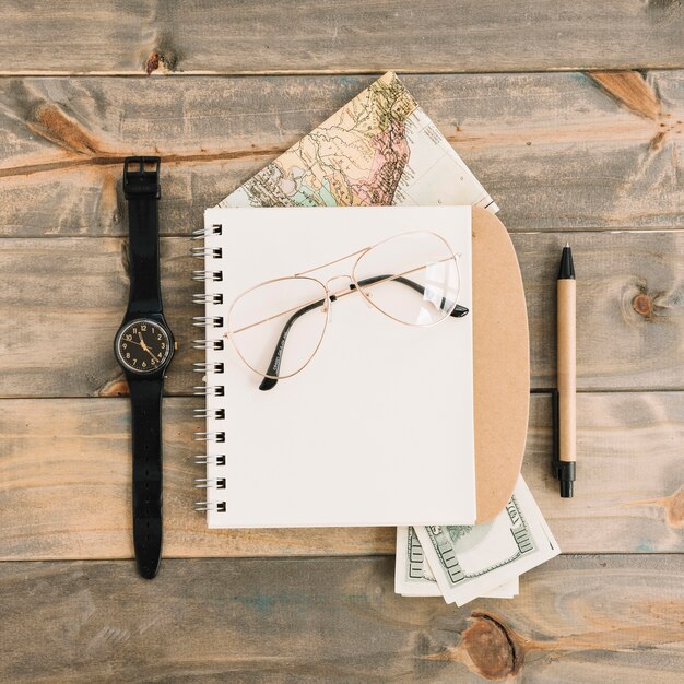 An overhead view of eyeglasses on spiral notepad; currency; map; wristwatch and pen on wooden plank background