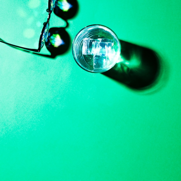 An overhead view of eyeglasses and water glass with shadow on green background