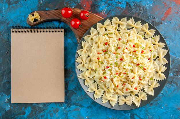 Vista dall'alto delle paste farfalle con verdure su un piatto nero pomodori su tagliere di legno e taccuino chiuso sul tavolo blu