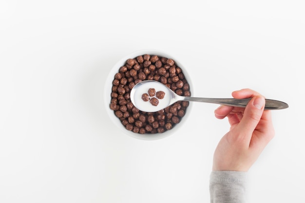 Free photo an overhead view of a female hand holding spoon with tasty cereal chocolate balls and milk in the bowl
