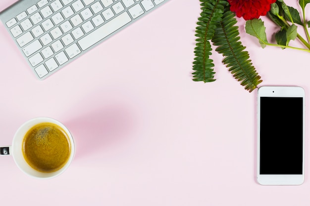 Free photo an overhead view of fern; red flower; keyboard; smartphone and cup of tea on pink background