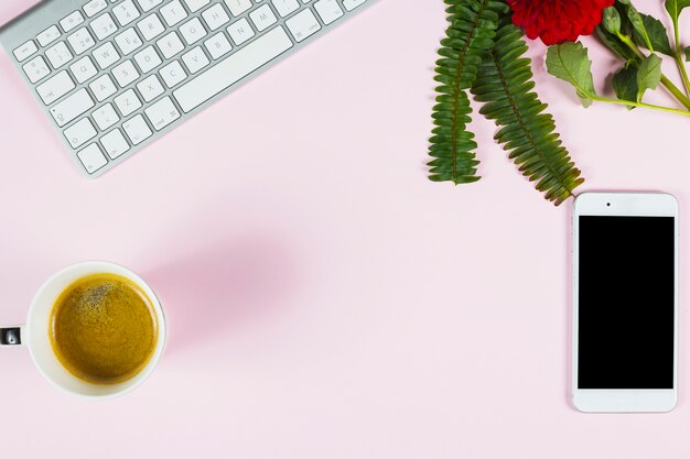 An overhead view of fern; red flower; keyboard; smartphone and cup of tea on pink background