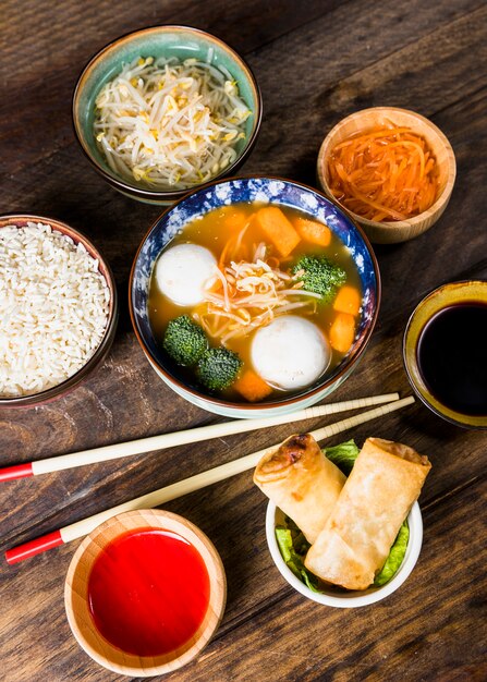 An overhead view of fish ball soup; rice; beans sprouts carrot and spring rolls with sauces and chopsticks over the table