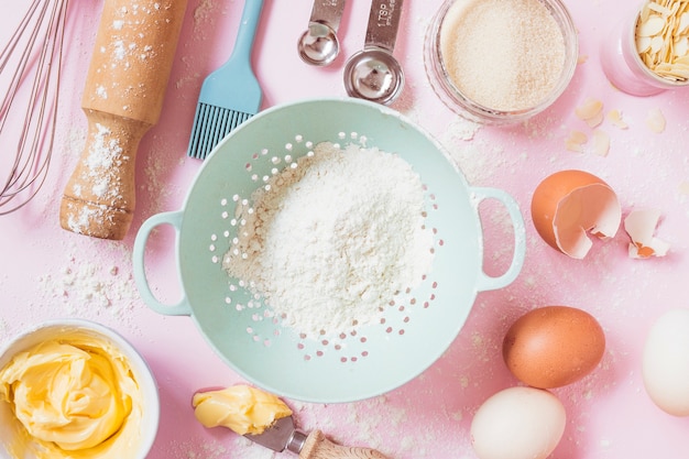 An overhead view of flour; eggs; butter and equipments on pink background