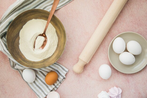 An overhead view of flour; eggs; rolling pin and napkin on colored backdrop