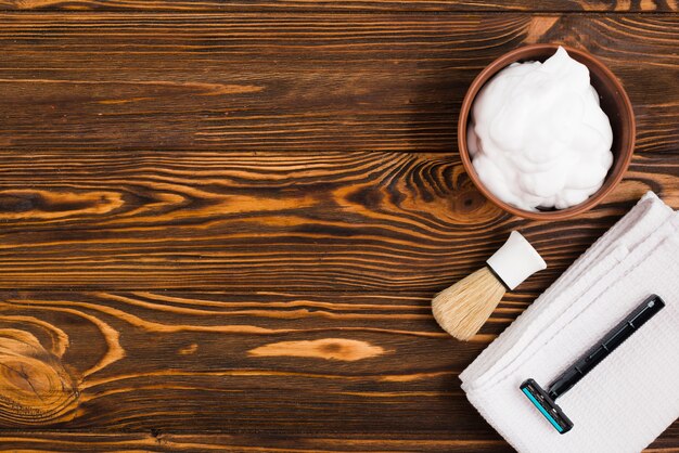An overhead view of foam bowl; shaving brush; razor and white folded napkin against wooden textured backdrop