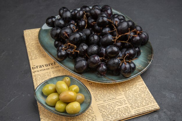 Вид сверху на свежие органические зеленые оливки и гроздья черного винограда на старой газете на темном фоне