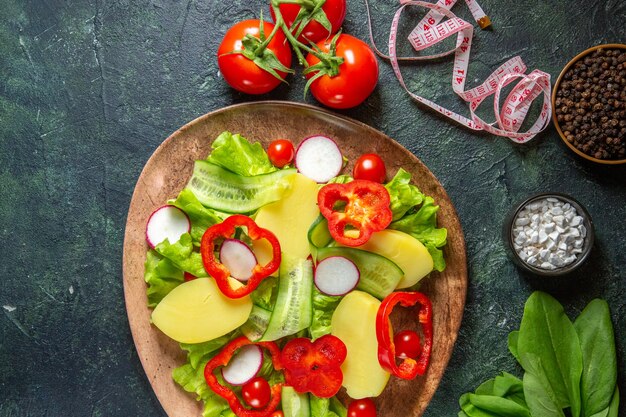 Overhead view of fresh peeled cut potatoes with red pepper radishes green tomatoes in a brown plate and meters spices on green black mix colors surface