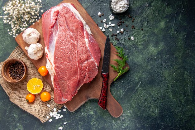 Overhead view of fresh raw red meats green garlics on brown wooden cutting board tomatoes pepper on nude color towel knife on dark color background